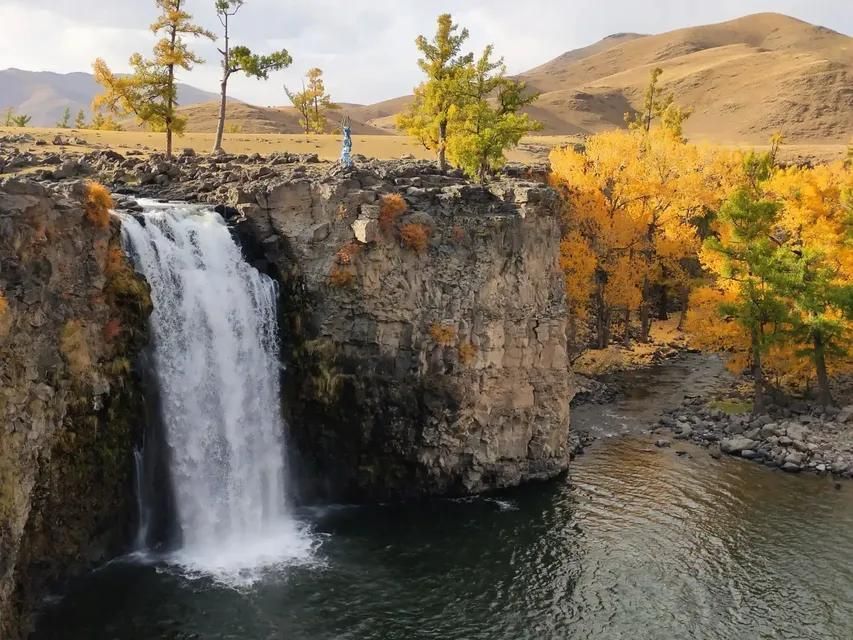A wide waterfall cascades over a rocky cliff into a river, surrounded by trees with yellow autumn leaves and rolling hills in the background.