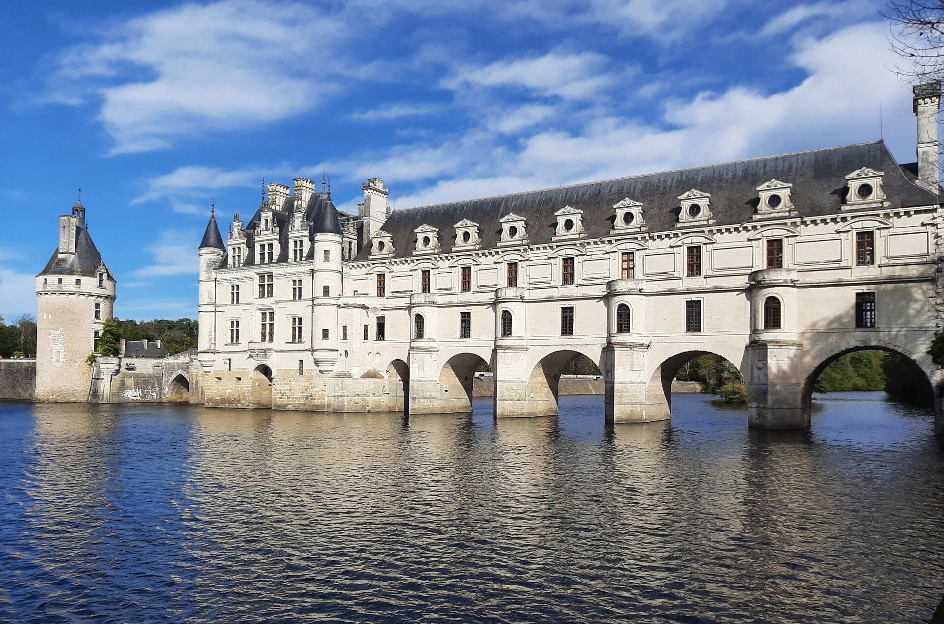 Un château de pierre blanche à tourelles bâti sur des fondations en arc enjambant une rivière, sous un ciel bleu avec quelques nuages.
