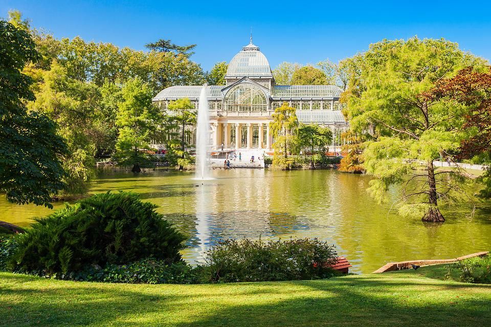 Un ornato palazzo di vetro si erge di fronte a un lago con una grande fontana, circondato da rigogliosi alberi verdi in un parco sotto un cielo azzurro e limpido.