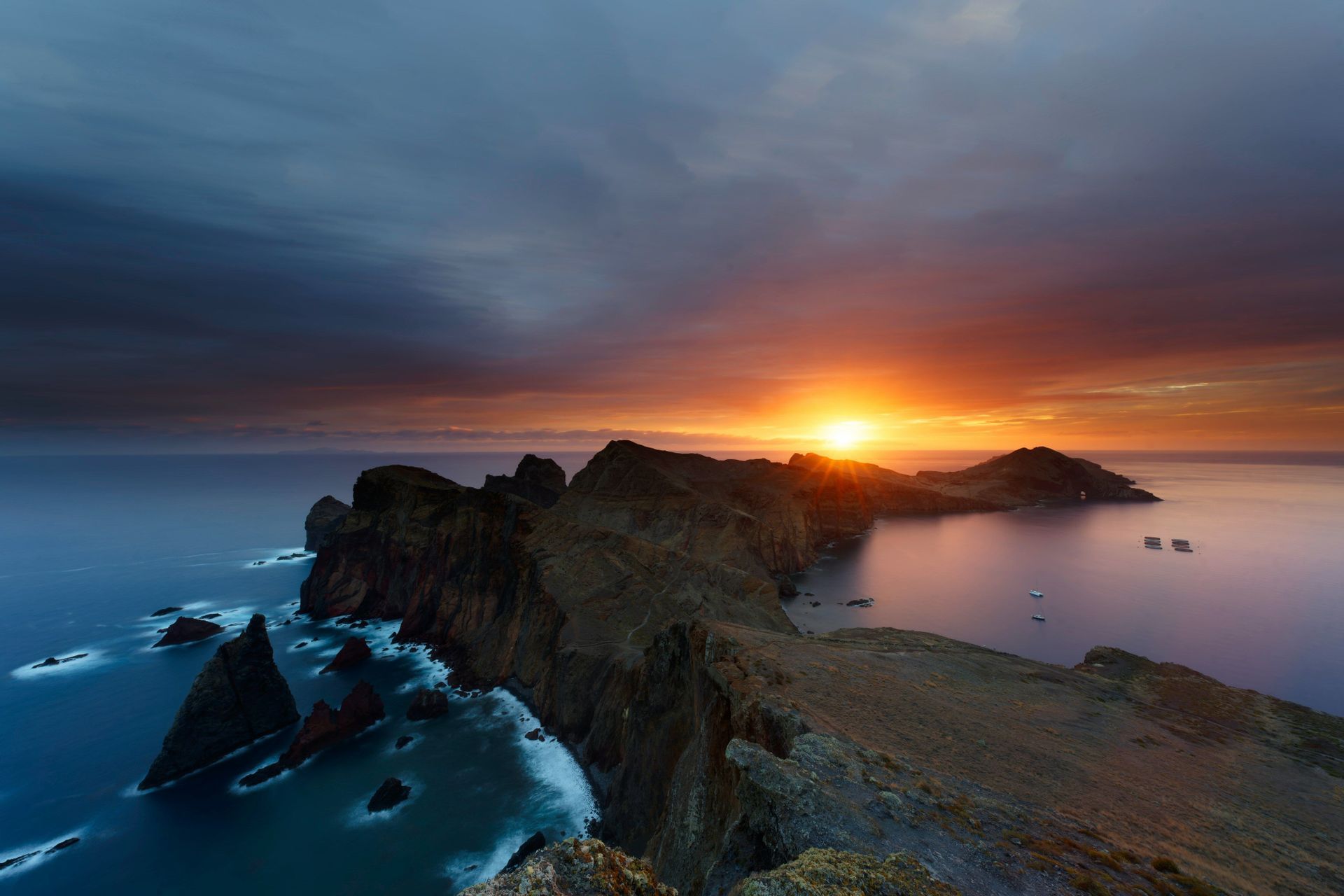 Una penisola rocciosa si estende nell'oceano sotto un cielo drammatico mentre il sole sorge sull'acqua.