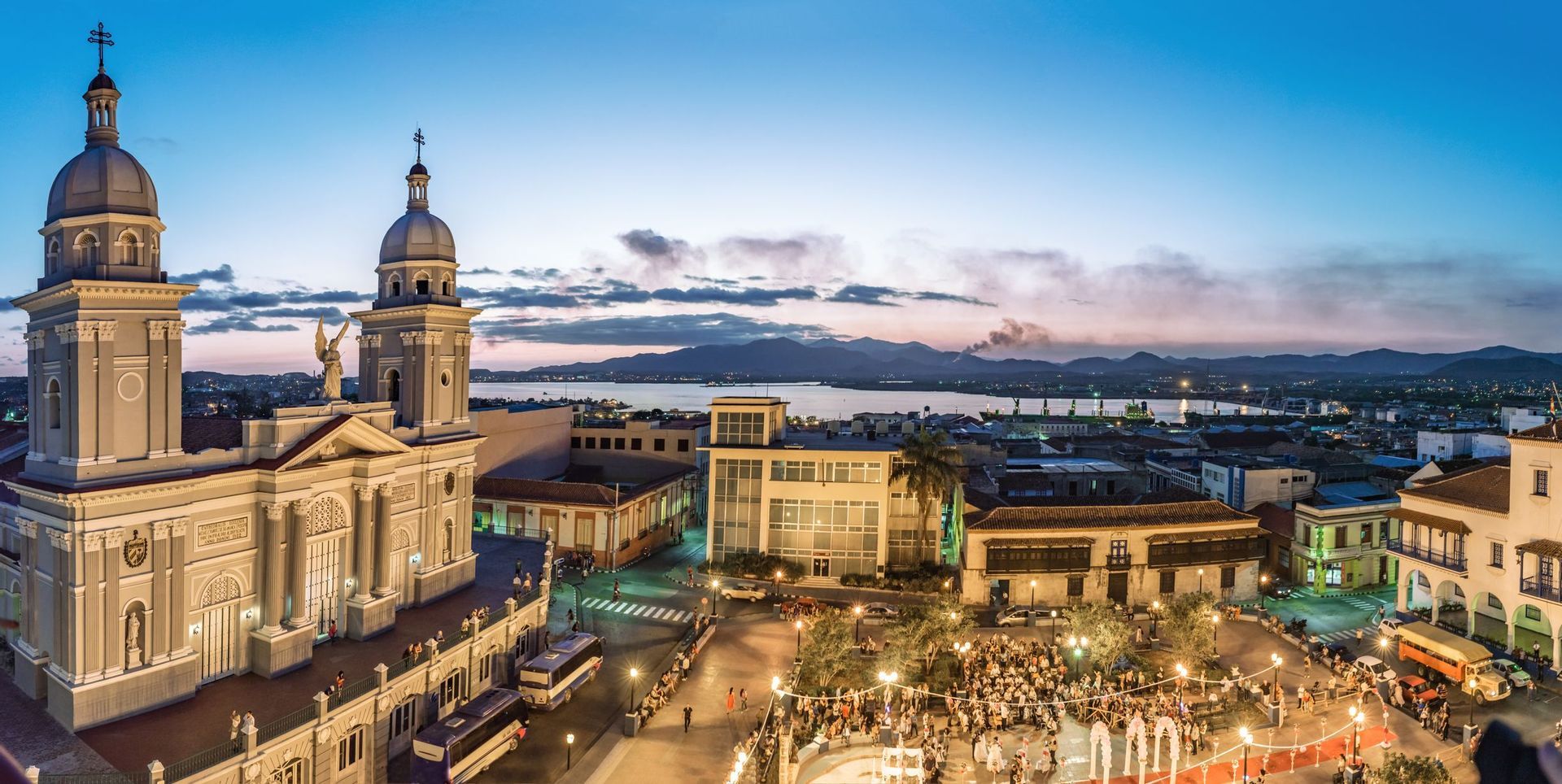 Una grande cattedrale con due campanili si affaccia su una vivace piazza cittadina al tramonto, con un porto e montagne sullo sfondo.