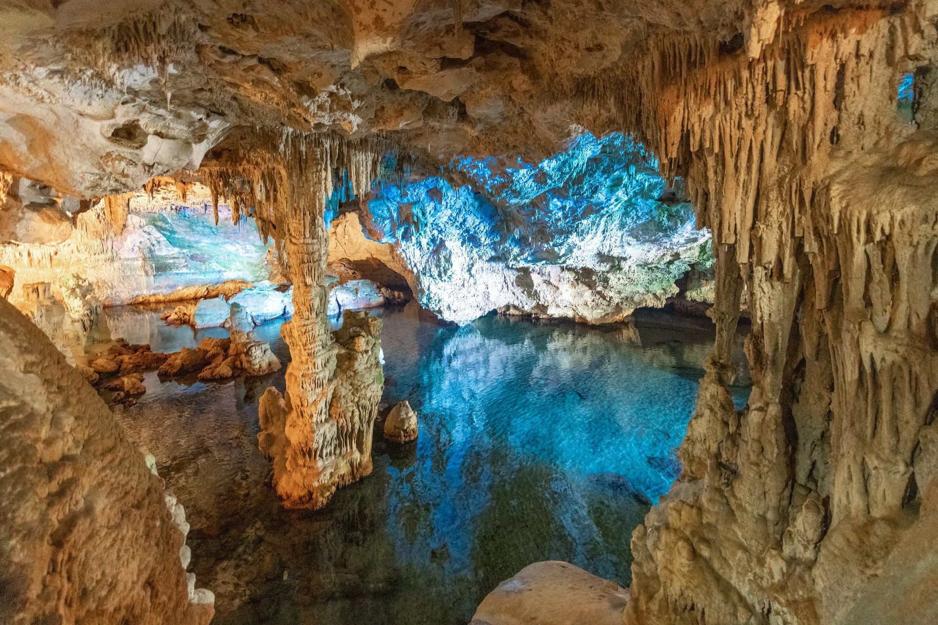 Stalattiti e stalagmiti all'interno di una vasta caverna con una limpida piscina sotterranea illuminata da luci blu e gialle.