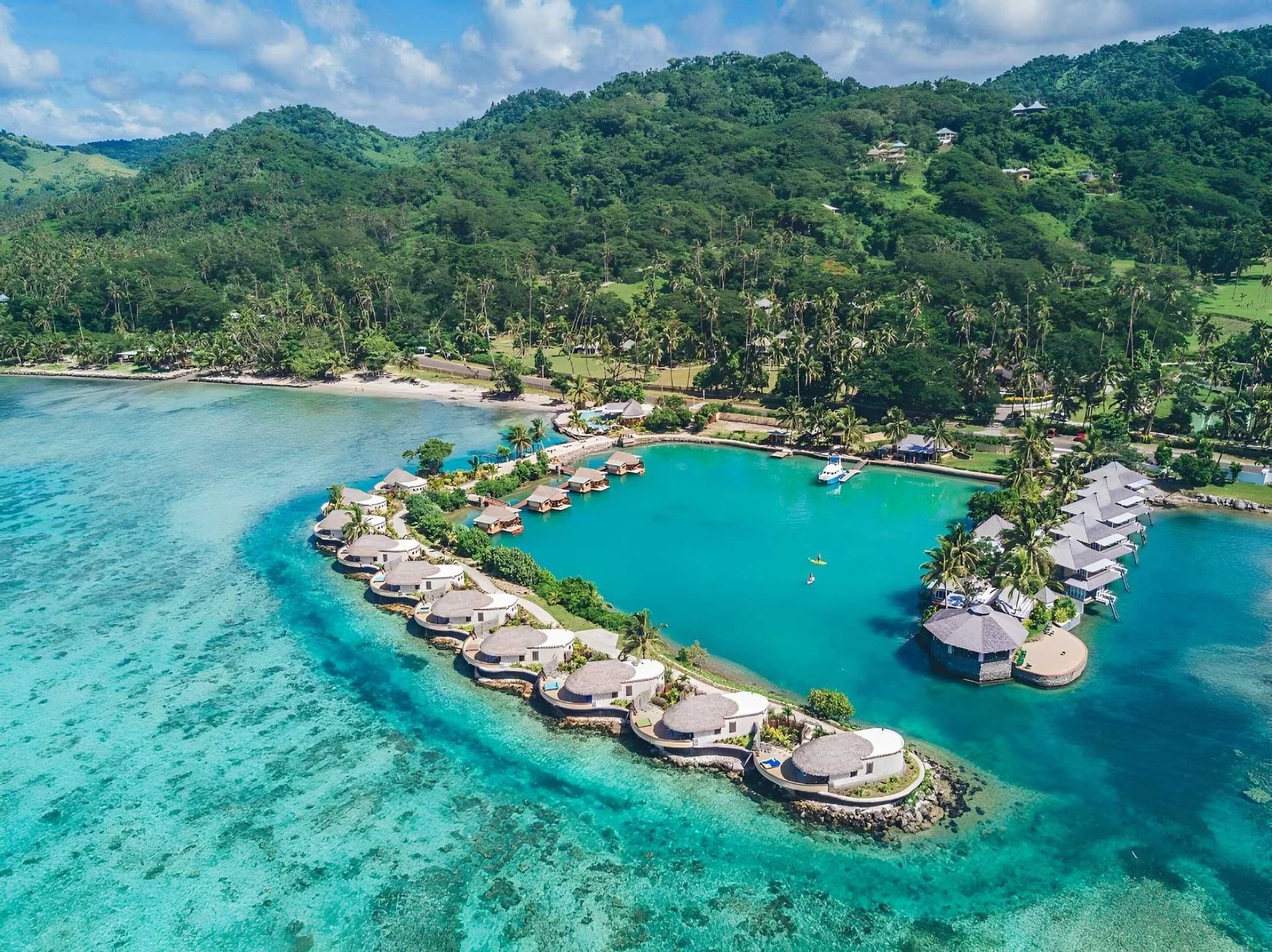 Vista aerea di bungalow sull'acqua che circondano una laguna turchese, con un'isola tropicale lussureggiante sullo sfondo.