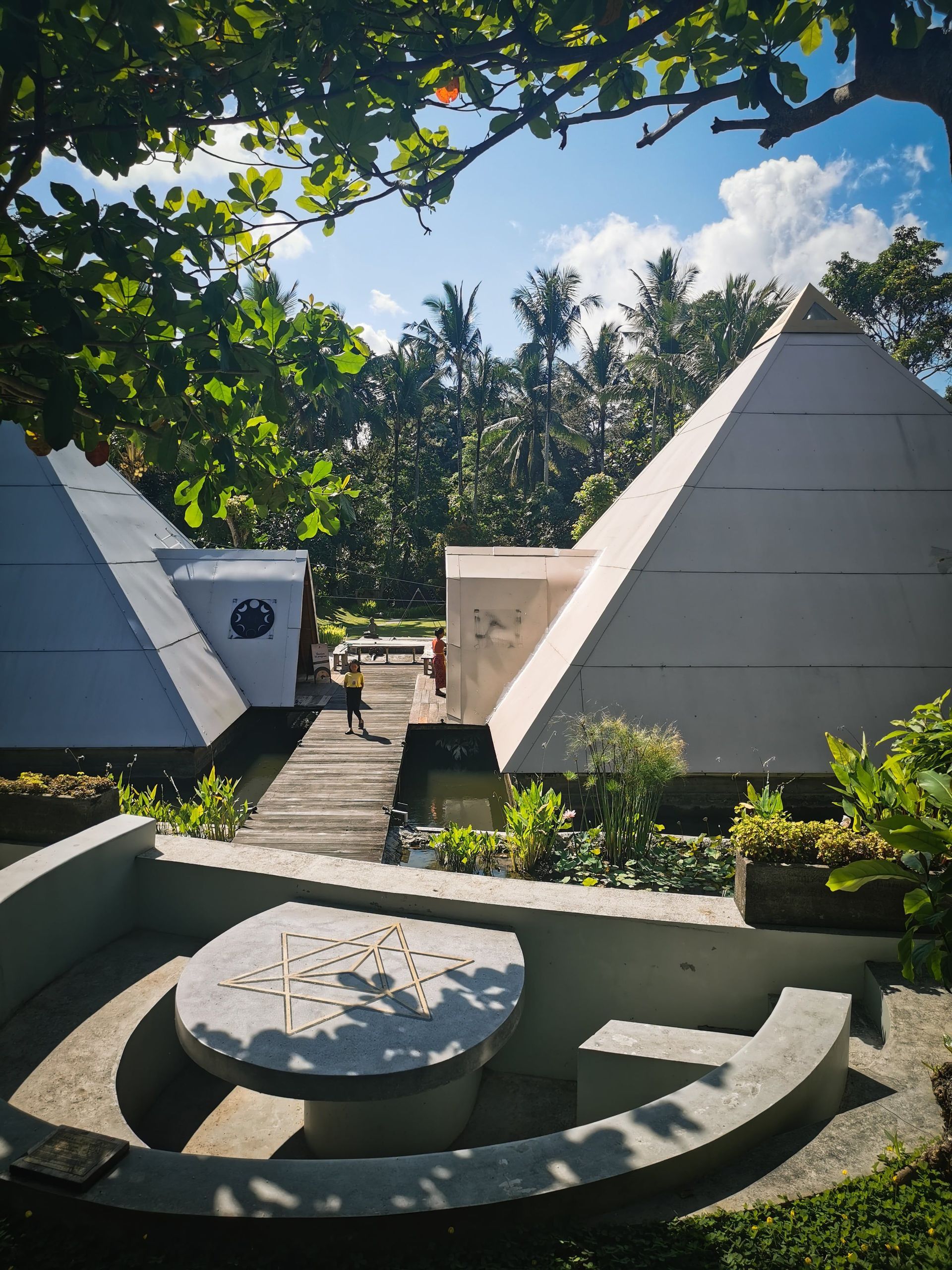 Ein sonniger Innenhof mit zwei großen weißen Pyramidenbauten, umgeben von üppiger tropischer Vegetation und einem kleinen Teich.