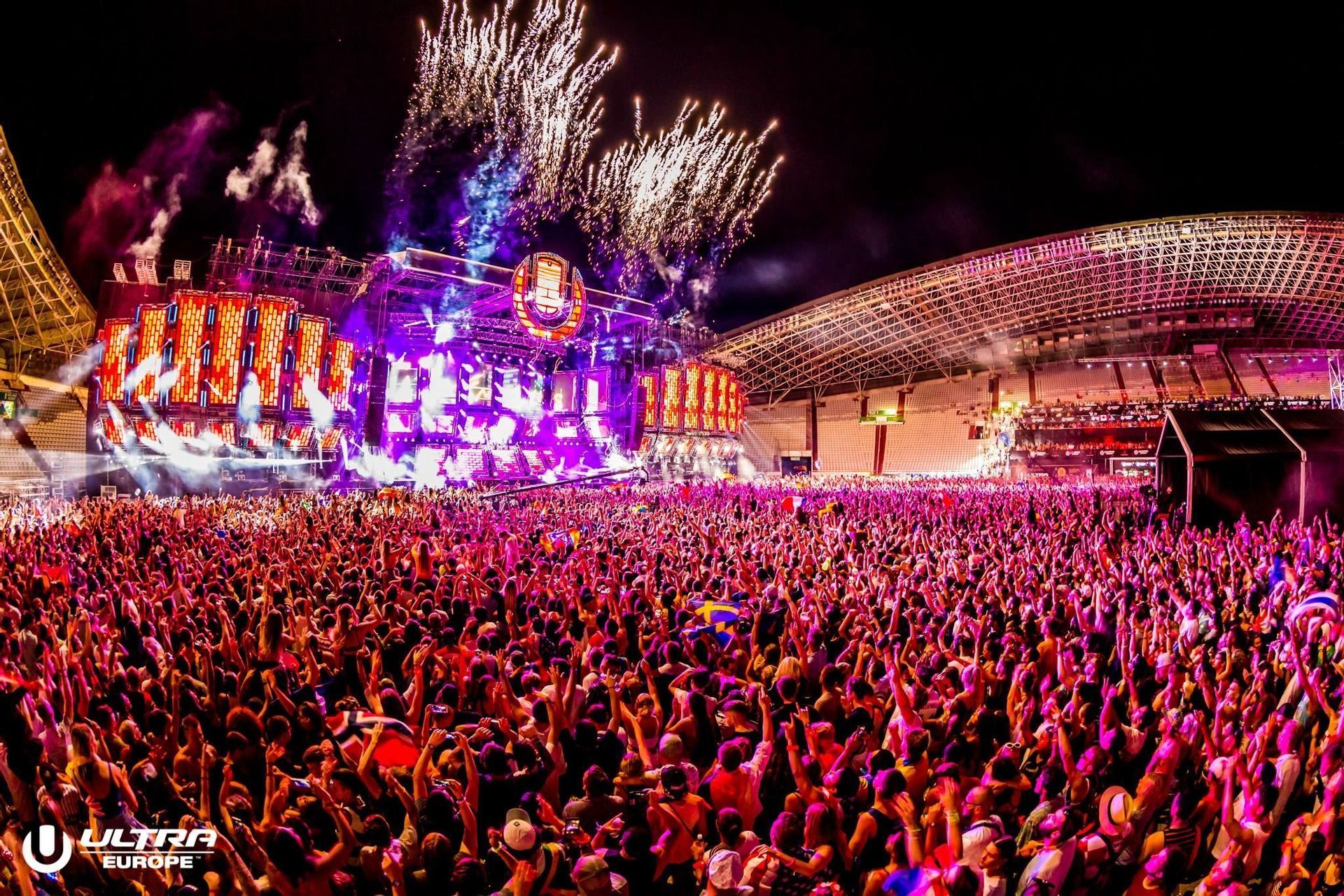 Una folla immensa con le mani alzate di fronte a un palco illuminato a giorno con fuochi d'artificio sopra al festival musicale Ultra Europe di notte.