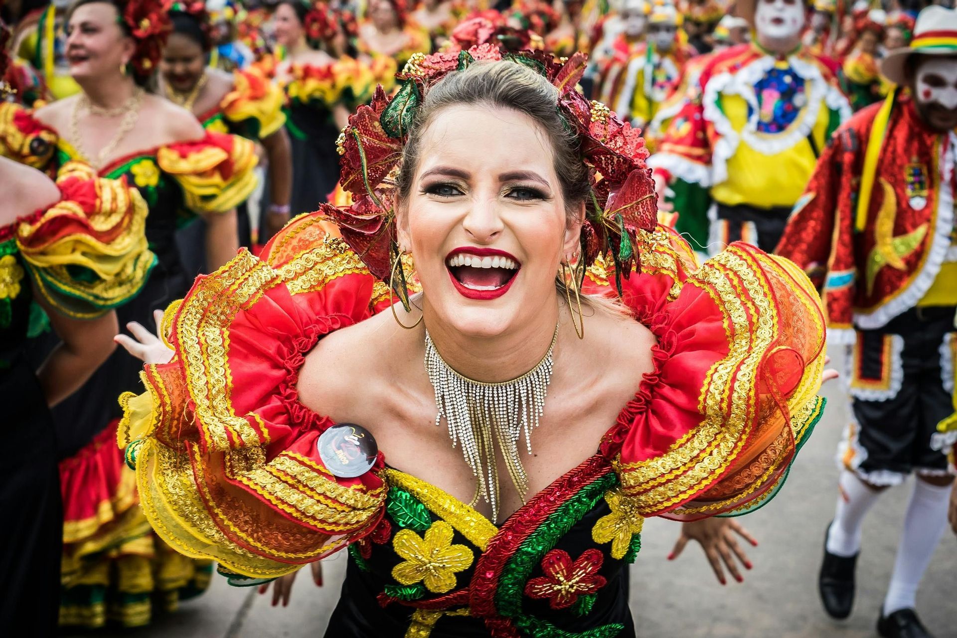 Una donna con un vivace costume rosso e giallo e maniche a balze sorride ampiamente durante una sfilata di carnevale.
