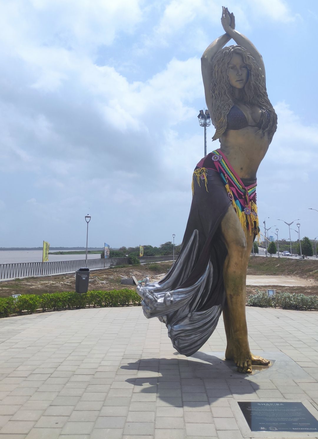 Un'alta statua di bronzo di Shakira in posa danzante si erge su una passeggiata lungofiume lastricata sotto un cielo nuvoloso.