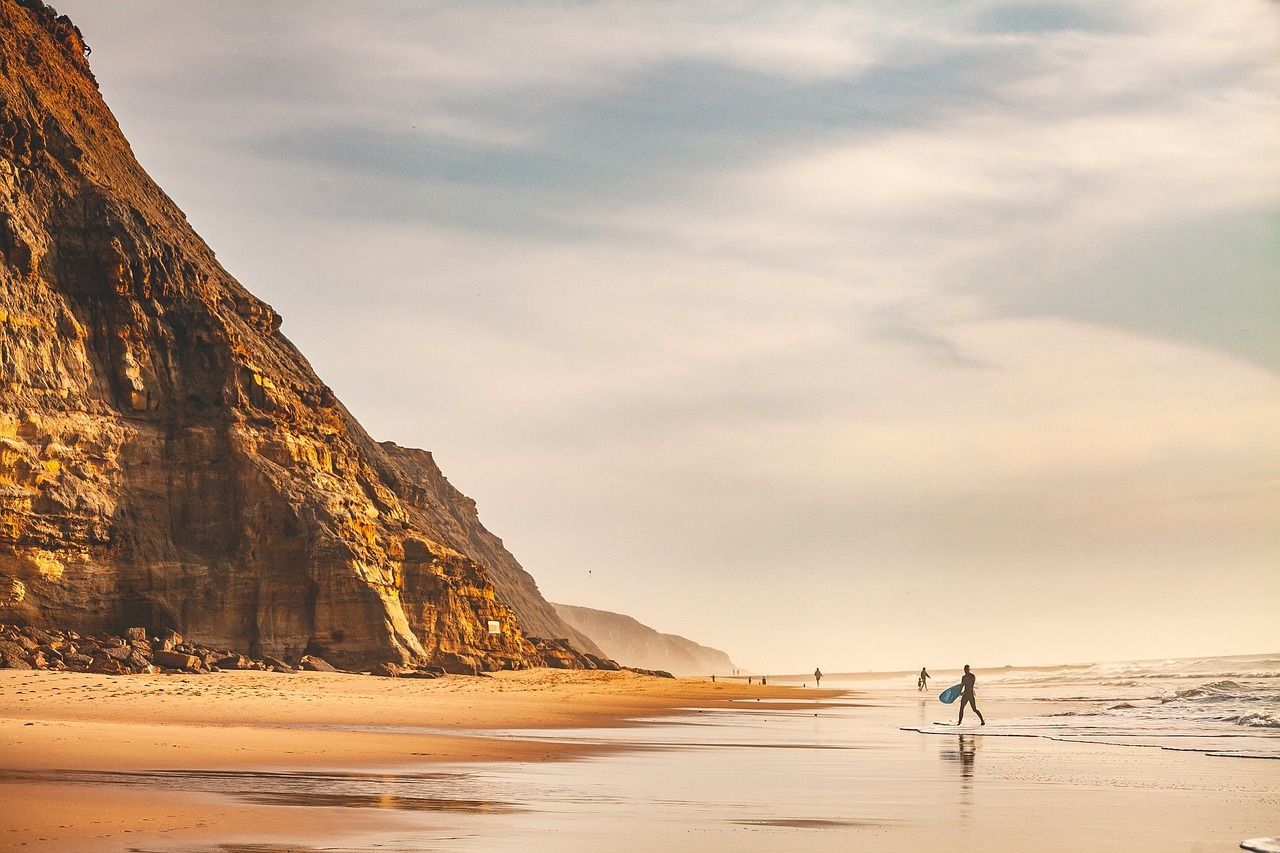 Ein Surfer mit Surfbrett geht am nassen Sand eines Strandes am Fuße einer großen, steilen Klippe entlang.