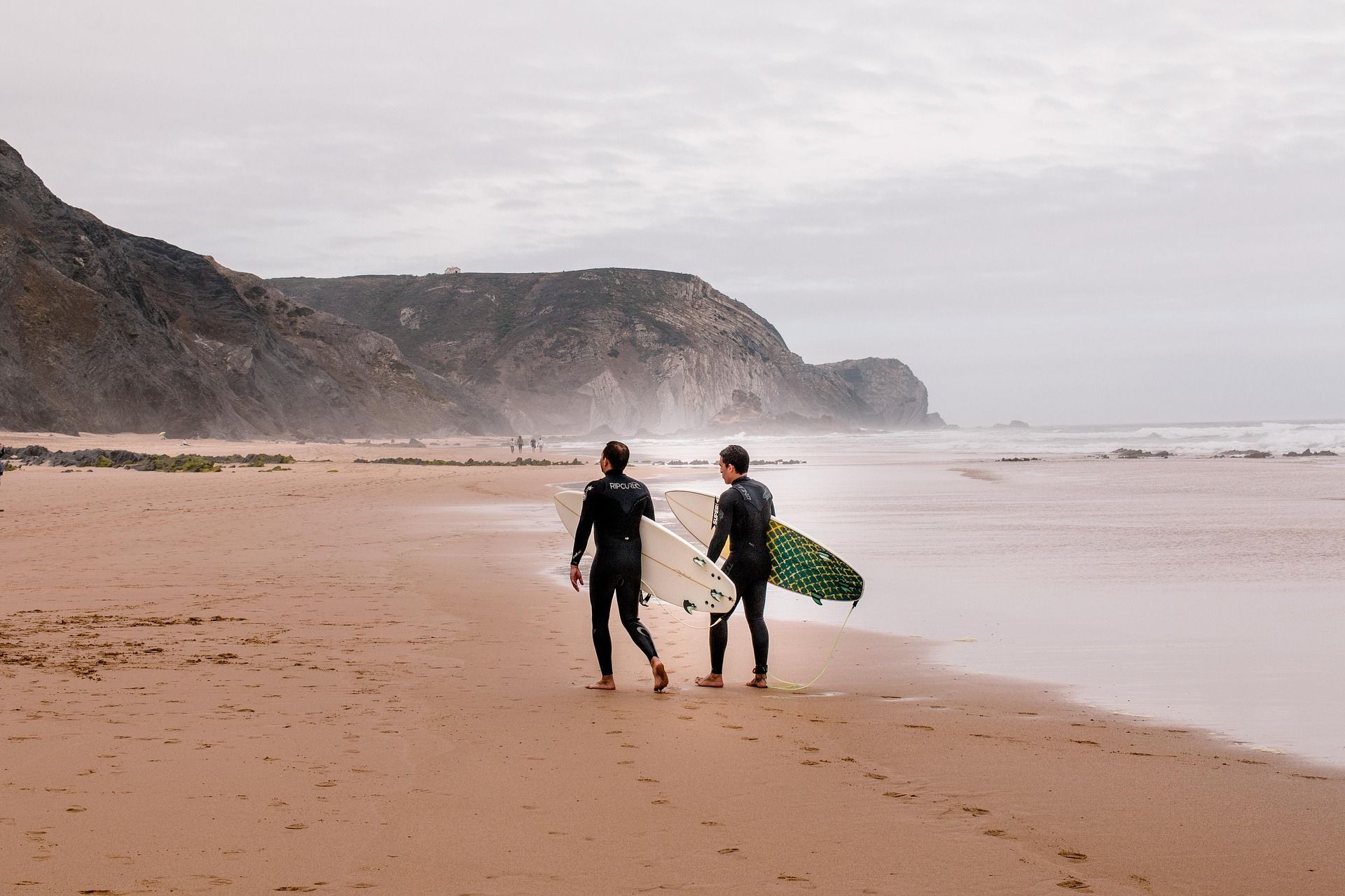 Zwei Surfer einer WeRoad-Gruppenreise in Neoprenanzügen tragen ihre Boards, während sie an einem Sandstrand auf den Ozean zugehen.