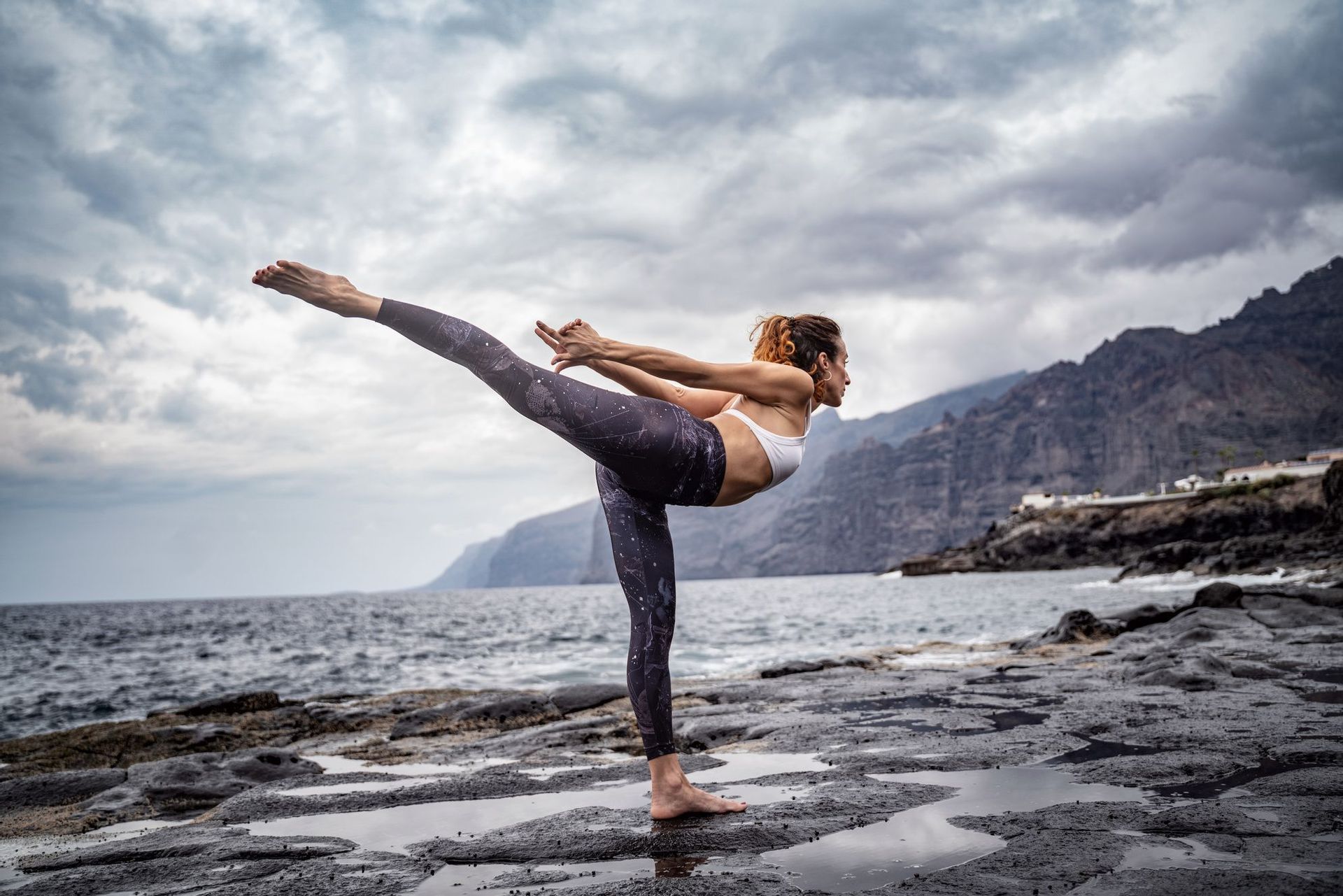 Una donna si bilancia in una posa yoga su una riva rocciosa e bagnata, con il mare e le montagne sotto un cielo nuvoloso.