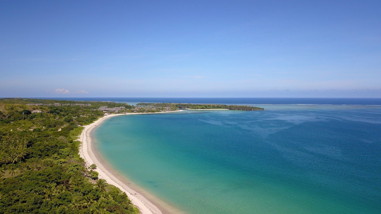 Veduta aerea di una spiaggia curva di sabbia bianca, che costeggia acqua turchese e una lussureggiante foresta verde, sotto un cielo sereno.