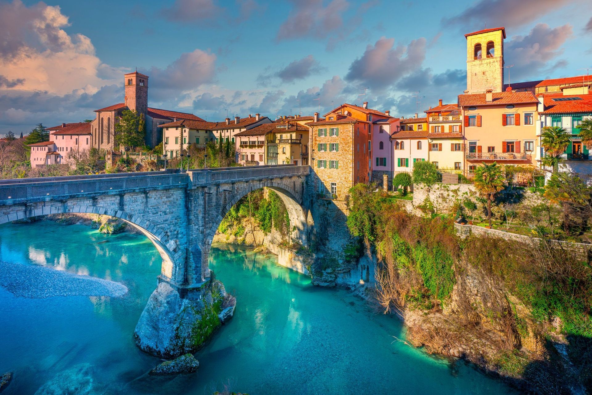 Un ponte ad arco in pietra attraversa un fiume turchese che conduce a un borgo colorato in collina con un campanile sotto un cielo parzialmente nuvoloso.