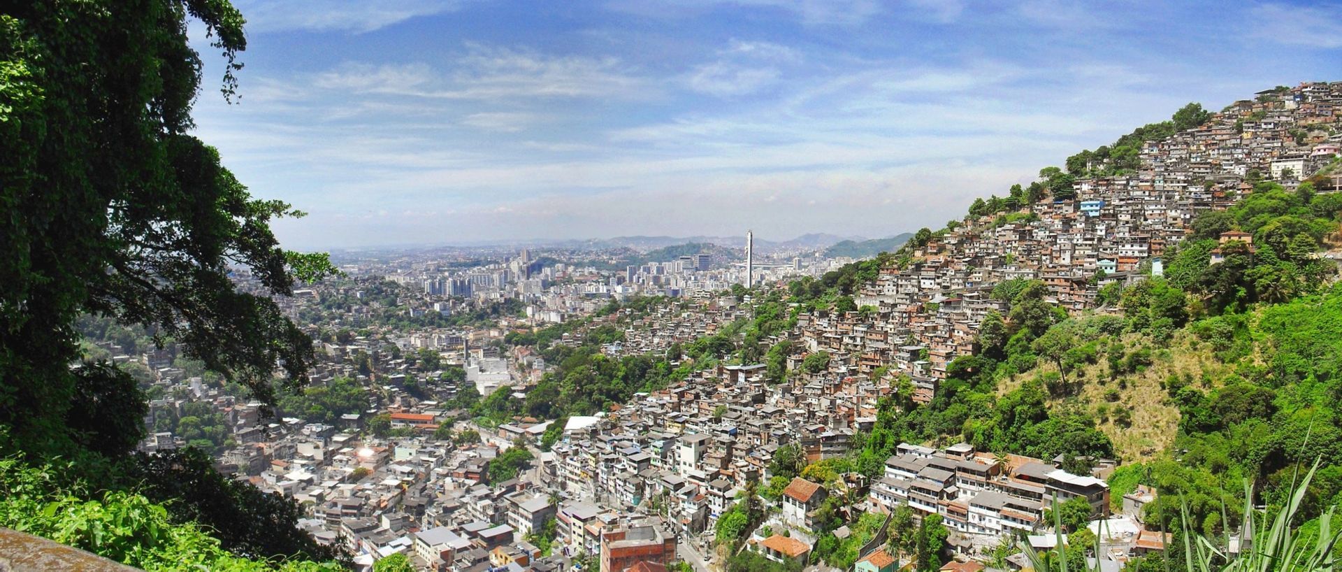 Una vista panoramica di una favela densamente popolata su una verde collina, con lo skyline della città in lontananza.