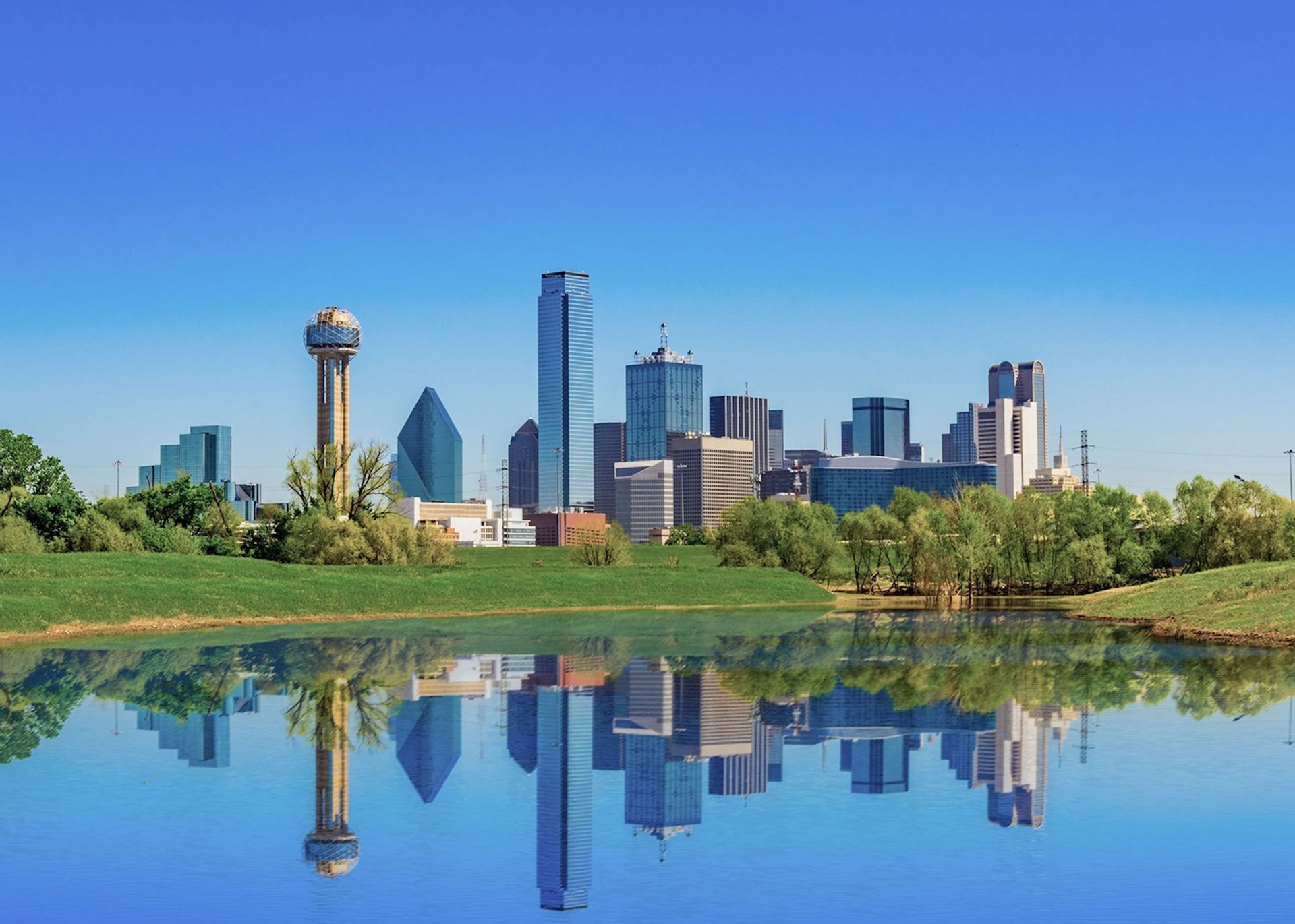 A modern city skyline with skyscrapers reflected perfectly in calm water, with a green grassy bank in the foreground.