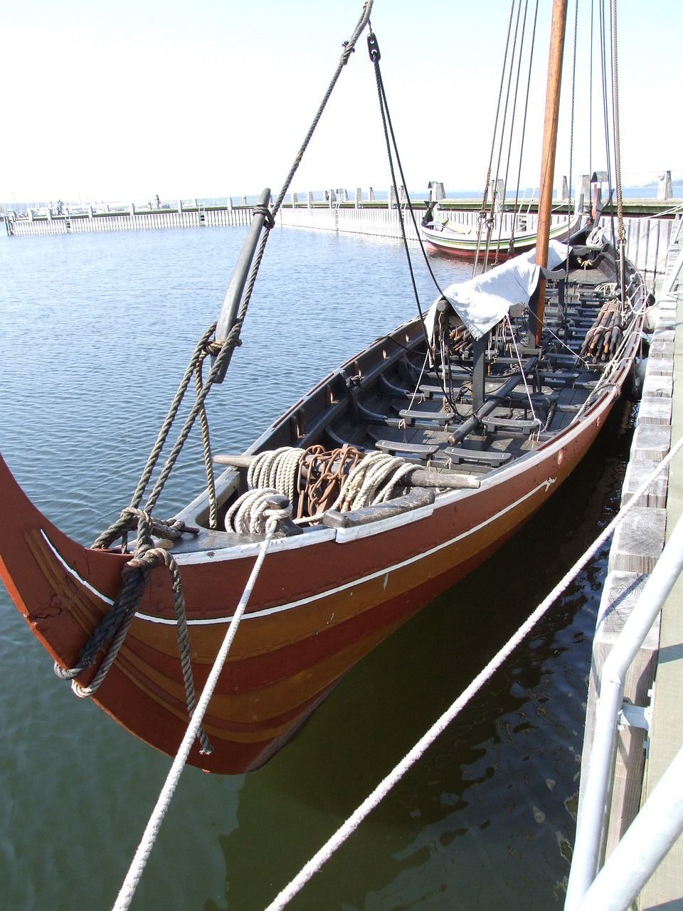 Eine hölzerne Wikinger-Langschiff-Replik mit aufgewickelten Seilen und einem Mast liegt an einem Holzsteg vertäut.