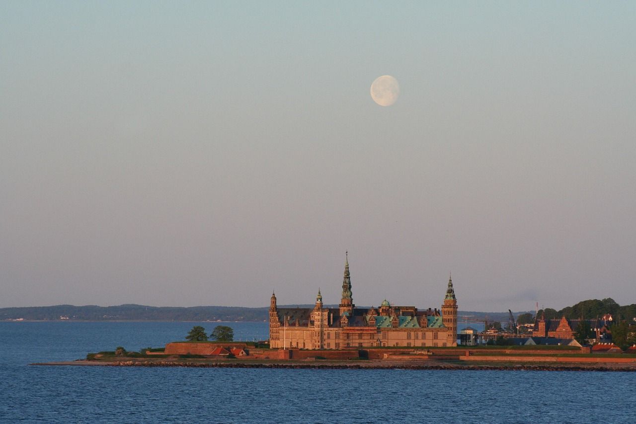 Ein großes Schloss mit spitzen Türmen thront auf einer Halbinsel am Meer, wobei ein Vollmond am Dämmerungshimmel sichtbar ist.