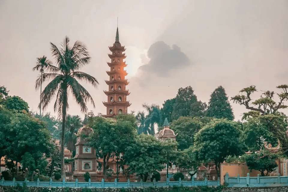 Une tour-pagode orange à plusieurs niveaux est entourée d'arbres verdoyants, y compris des palmiers, sous un ciel voilé au coucher du soleil.