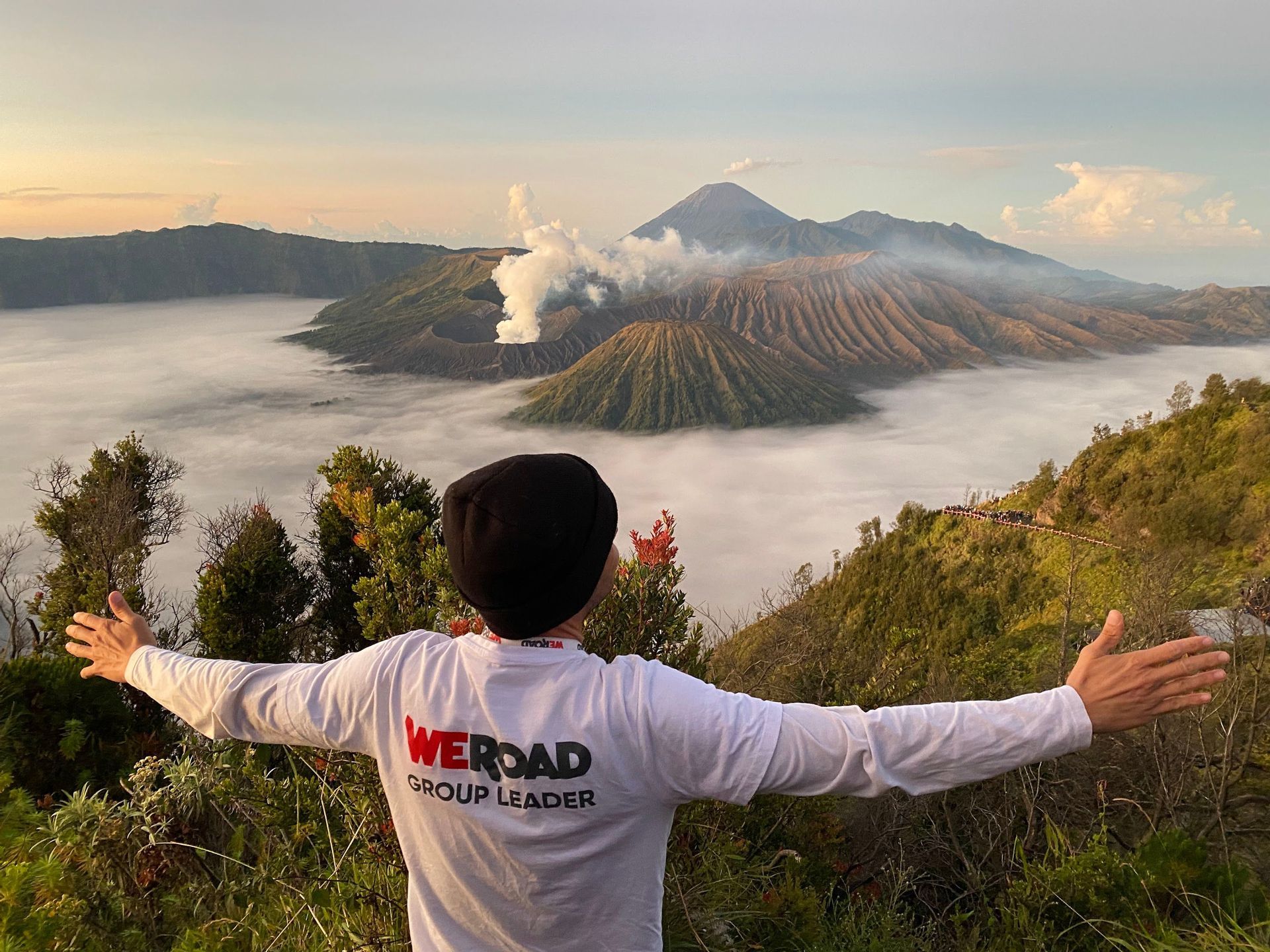 Un chef de groupe WeRoad, vu de dos, se tient les bras tendus, surplombant une vallée de nuages et des cratères volcaniques fumants à l'aube.