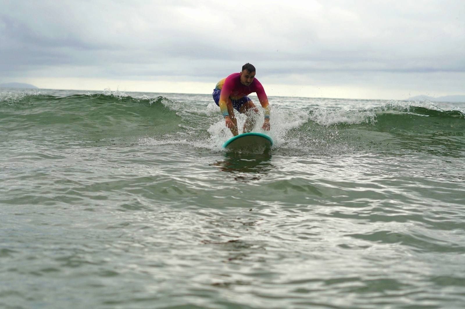 Un hombre con una camiseta de licra colorida surfea una pequeña ola en el océano bajo un cielo nublado.