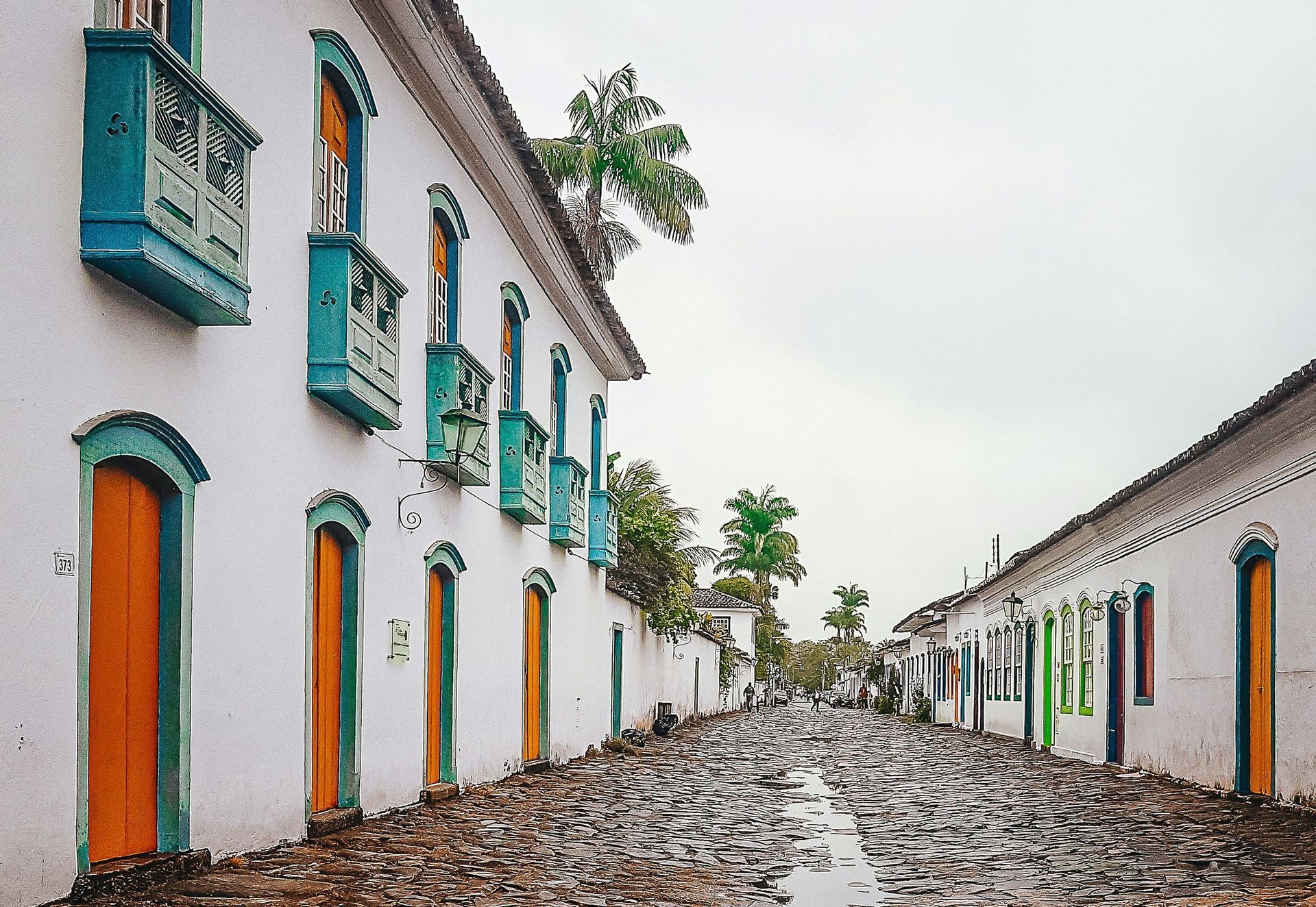 Una calle adoquinada mojada flanqueada por edificios coloniales encalados con puertas, ventanas y balcones coloridos bajo un cielo cubierto.