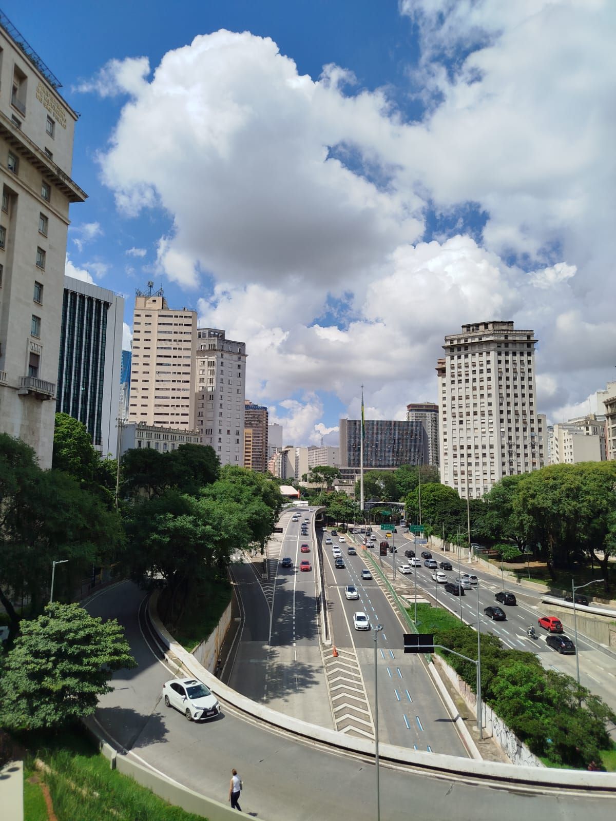 Vista elevada de una autopista de varios carriles que atraviesa una ciudad con rascacielos y exuberantes árboles bajo un cielo azul nublado.