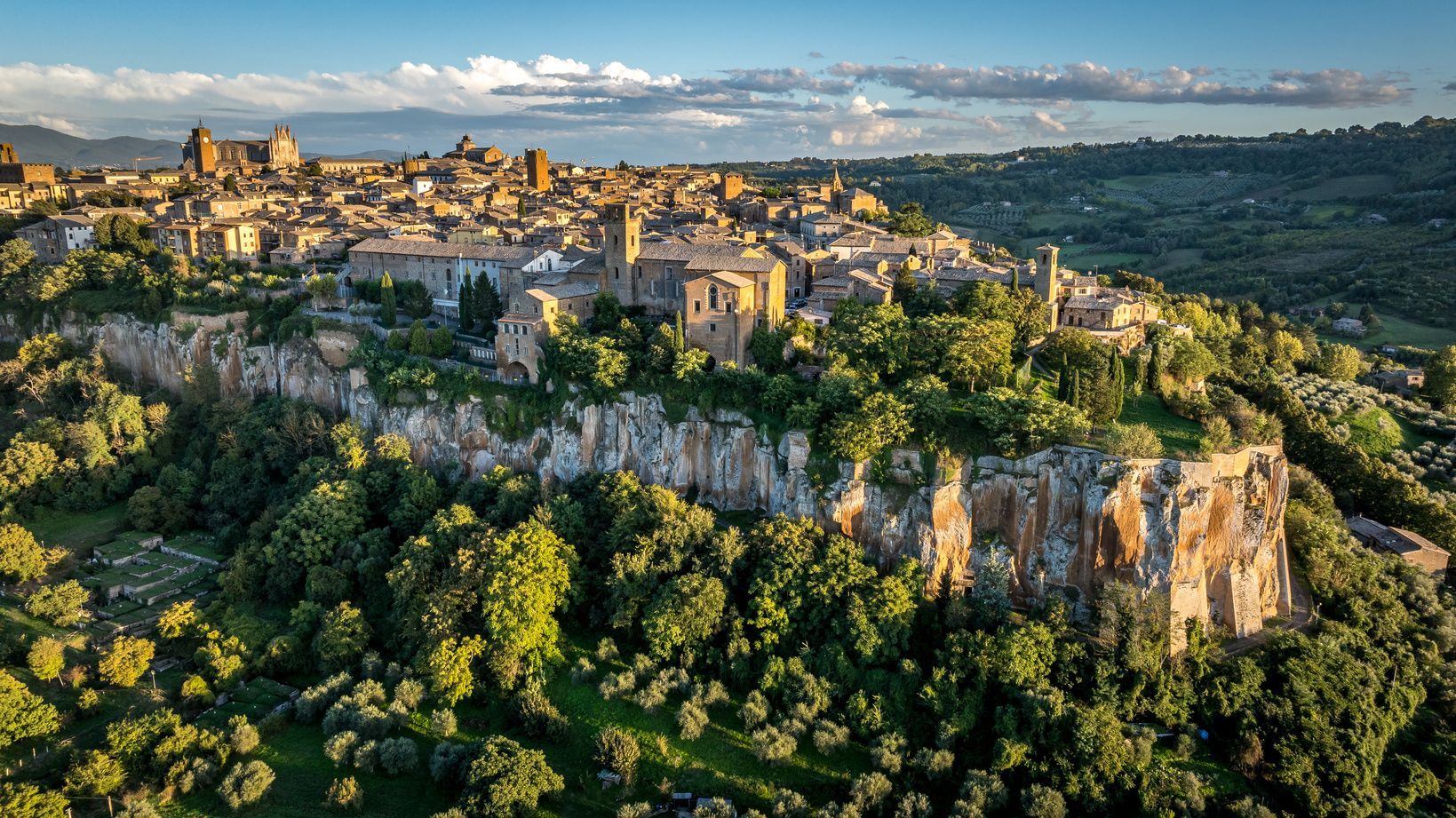 A historic town with stone buildings sits atop a steep, rocky cliff surrounded by dense green forests and rolling hills.