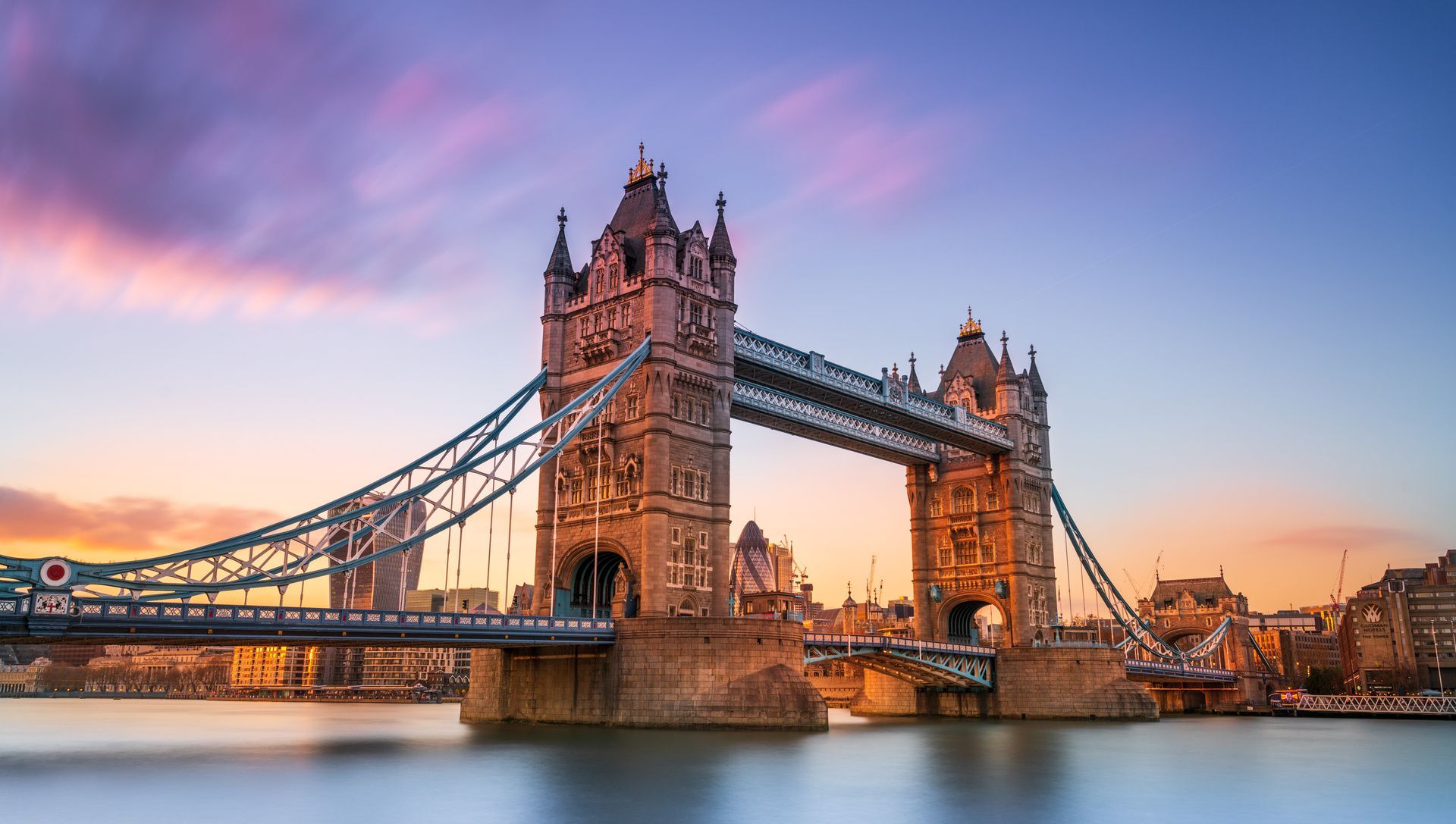 A long-exposure shot of the Tower Bridge in London spanning the River Thames at sunset, under a purple and orange sky.