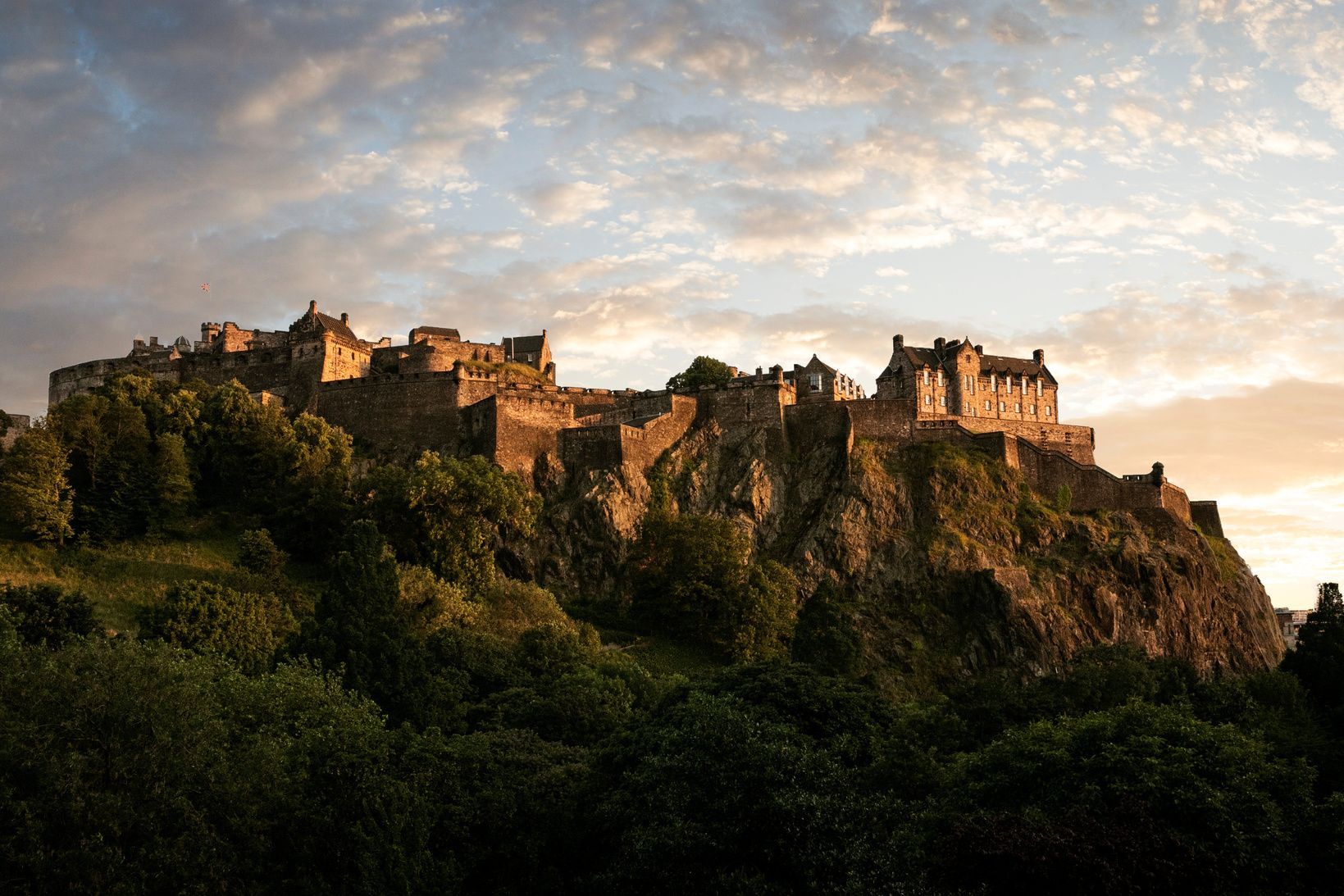 A large stone castle sits atop a rocky, tree-covered hill, illuminated by sunset against a cloudy sky.