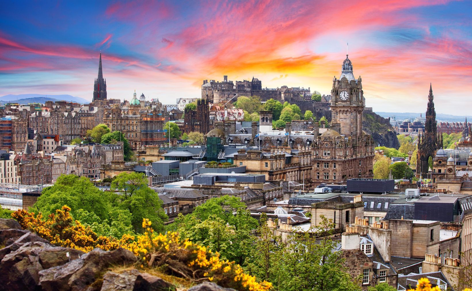 A historic European cityscape featuring a castle and clock towers, viewed from a hill with yellow flowers under a colorful sunset sky.