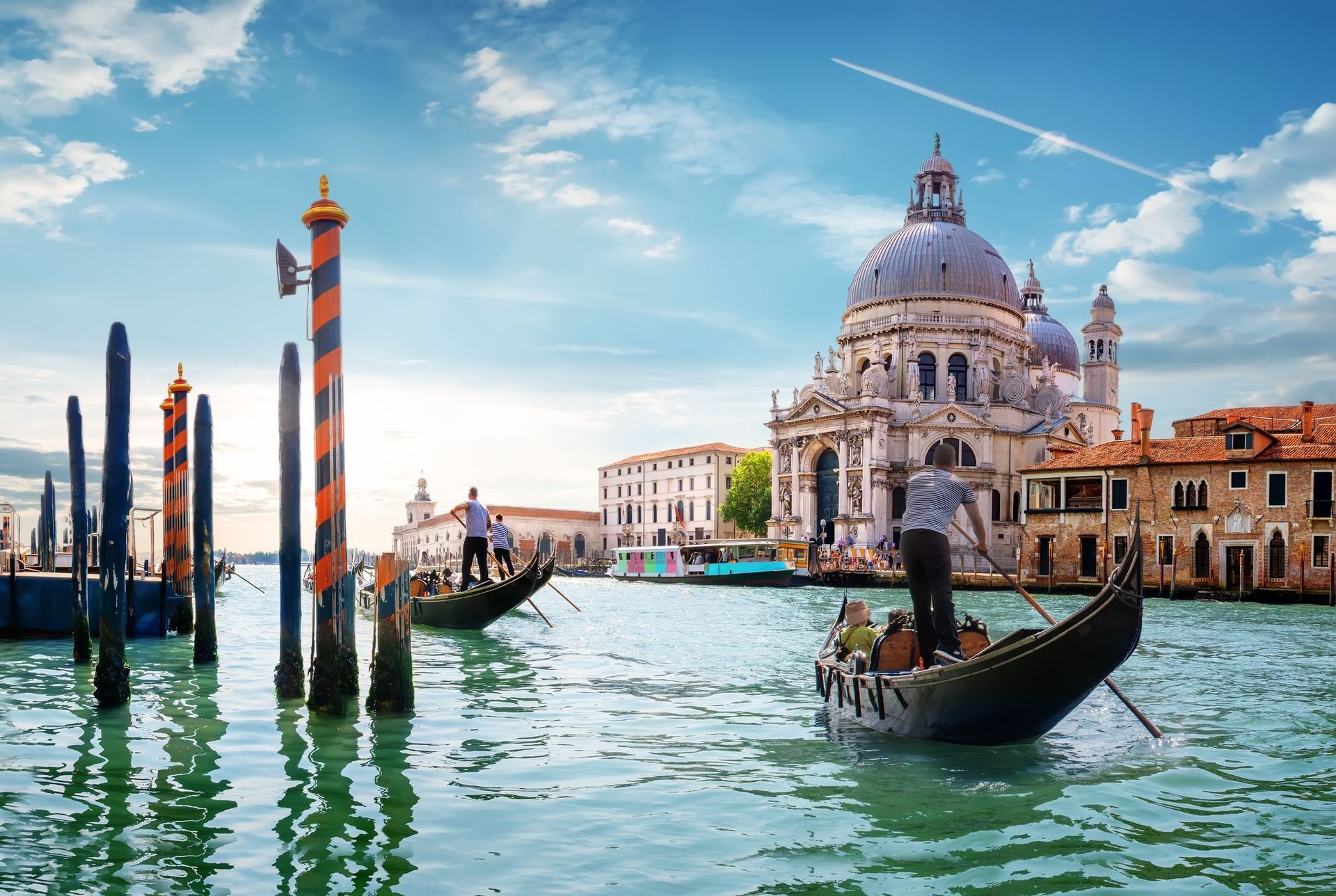 Two gondolas with passengers and gondoliers navigate a wide canal in front of a historic domed basilica under a partly cloudy sky.