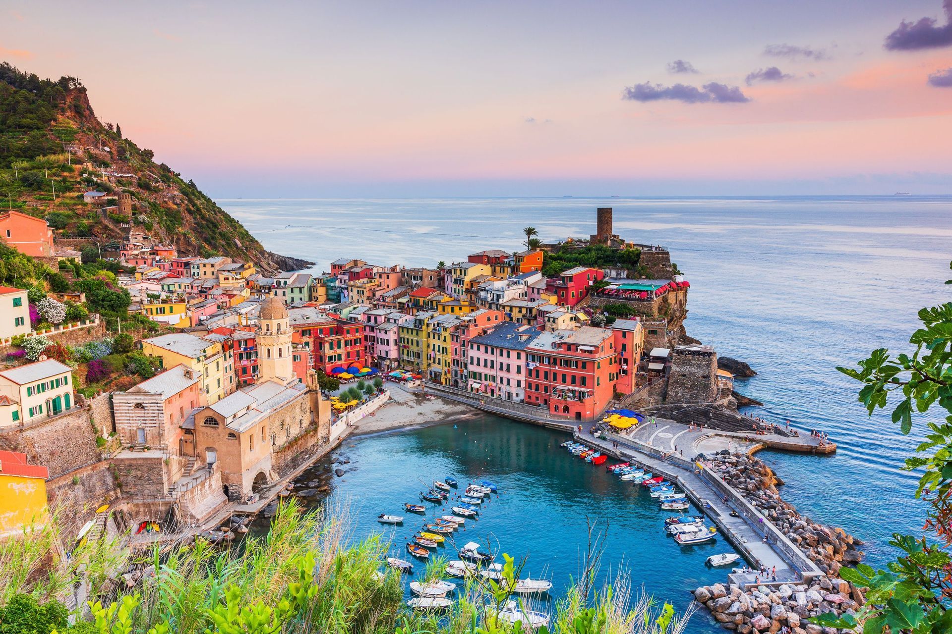 A colorful village on a steep cliff overlooks a small harbor filled with boats as the sun sets over the sea.