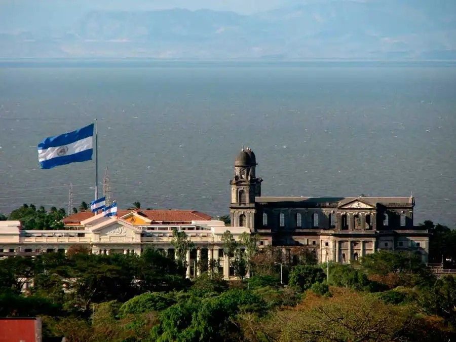 La bandiera nicaraguense sventola accanto a un edificio classico e una cattedrale su una collina lussureggiante che domina un grande lago.