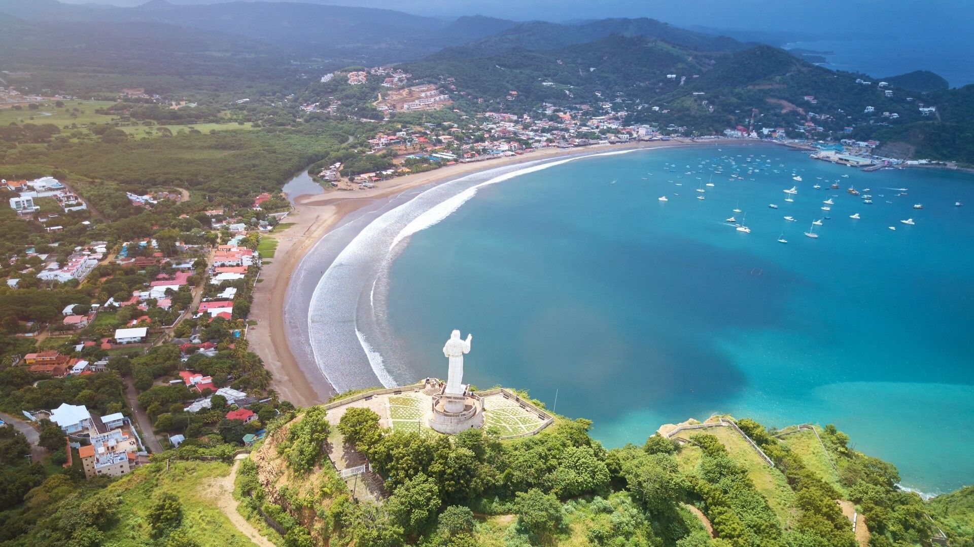 Una vista aerea di una grande statua bianca su una collina che domina una baia a forma di mezzaluna con una spiaggia, una città costiera e barche sull'acqua.