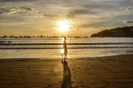 Una persona cammina in controluce lungo la riva al tramonto, con la sua lunga ombra proiettata sulla sabbia bagnata.