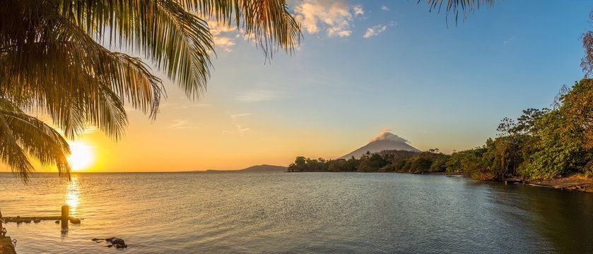 Il sole tramonta su uno specchio d'acqua calmo con un vulcano in lontananza, incorniciato da foglie di palma.