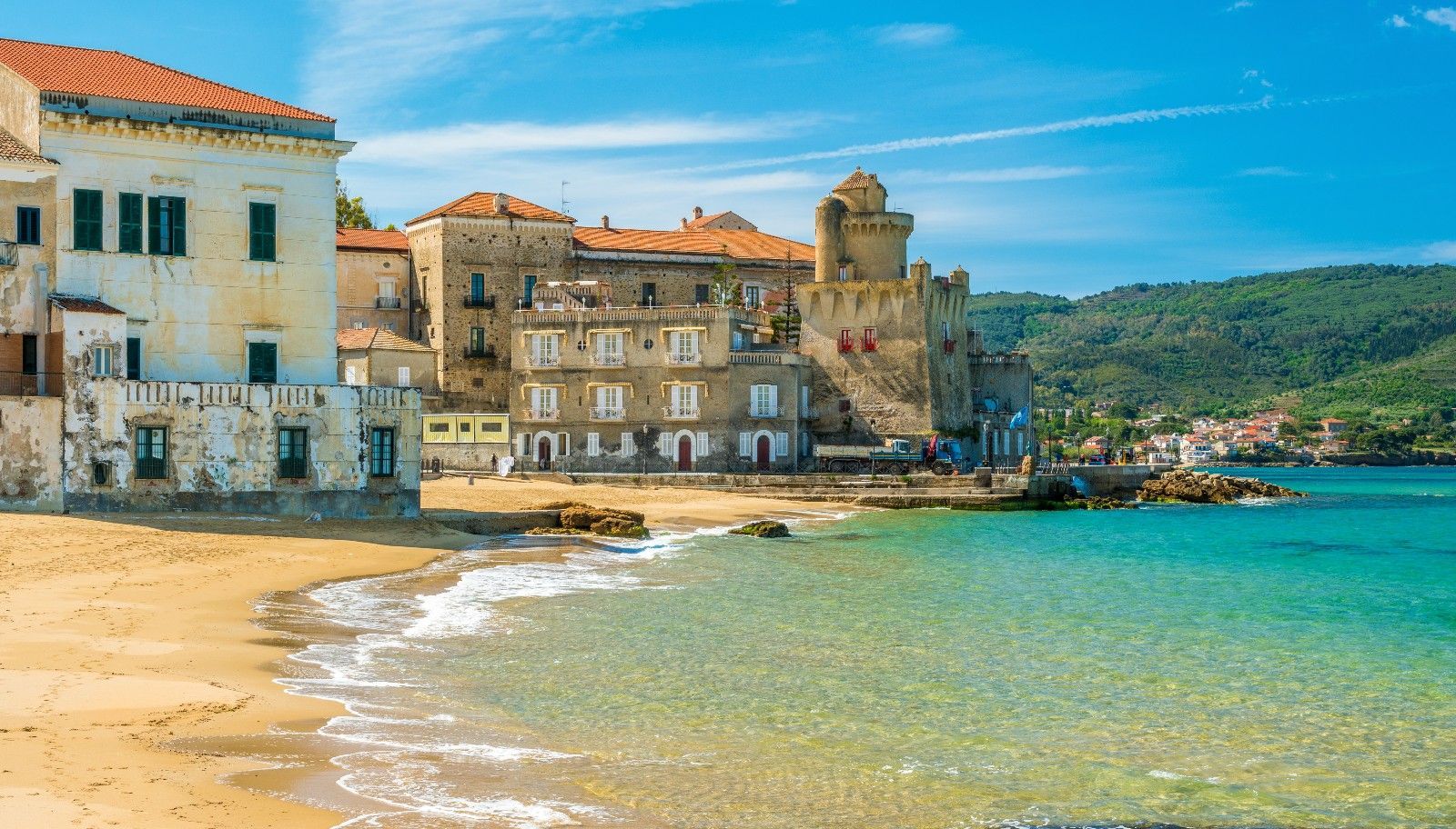 Un pueblo costero con edificios históricos y un castillo se asienta en una playa de arena junto a aguas cristalinas de color turquesa bajo un cielo azul.