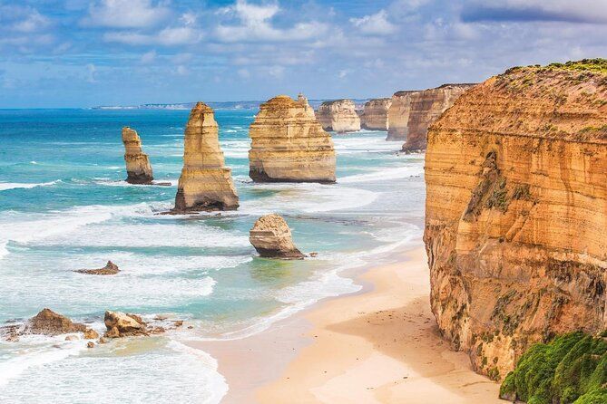 Sonnenbeschienene Kalksteinfelsen ragen aus dem türkisfarbenen Meer, umrahmt von einem Sandstrand und steilen Klippen unter einem teilweise bewölkten Himmel.
