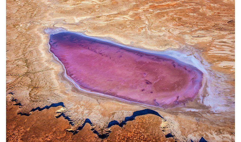 Eine Luftaufnahme eines länglichen rosa Sees, umgeben von einer trockenen, kargen Landschaft in Braun- und Orangetönen.