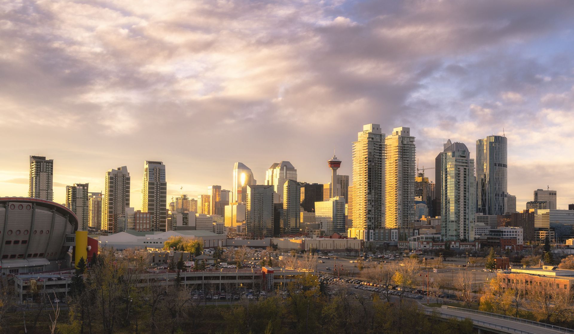 A cityscape with modern skyscrapers and a large domed stadium bathed in the golden light of sunset under a cloudy sky.