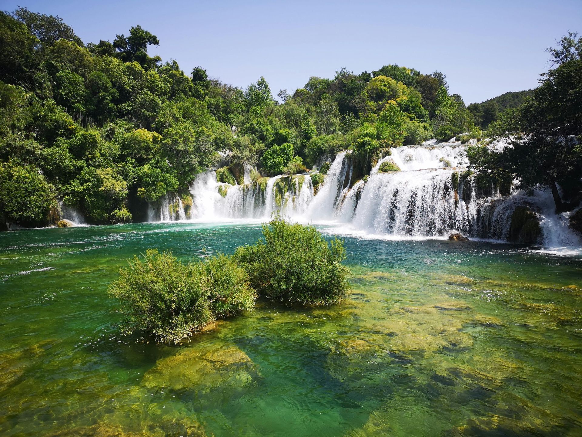 Mehrere breite Wasserfälle stürzen in einen klaren, türkisen Fluss, umgeben von einem dichten grünen Wald unter klarem Himmel.