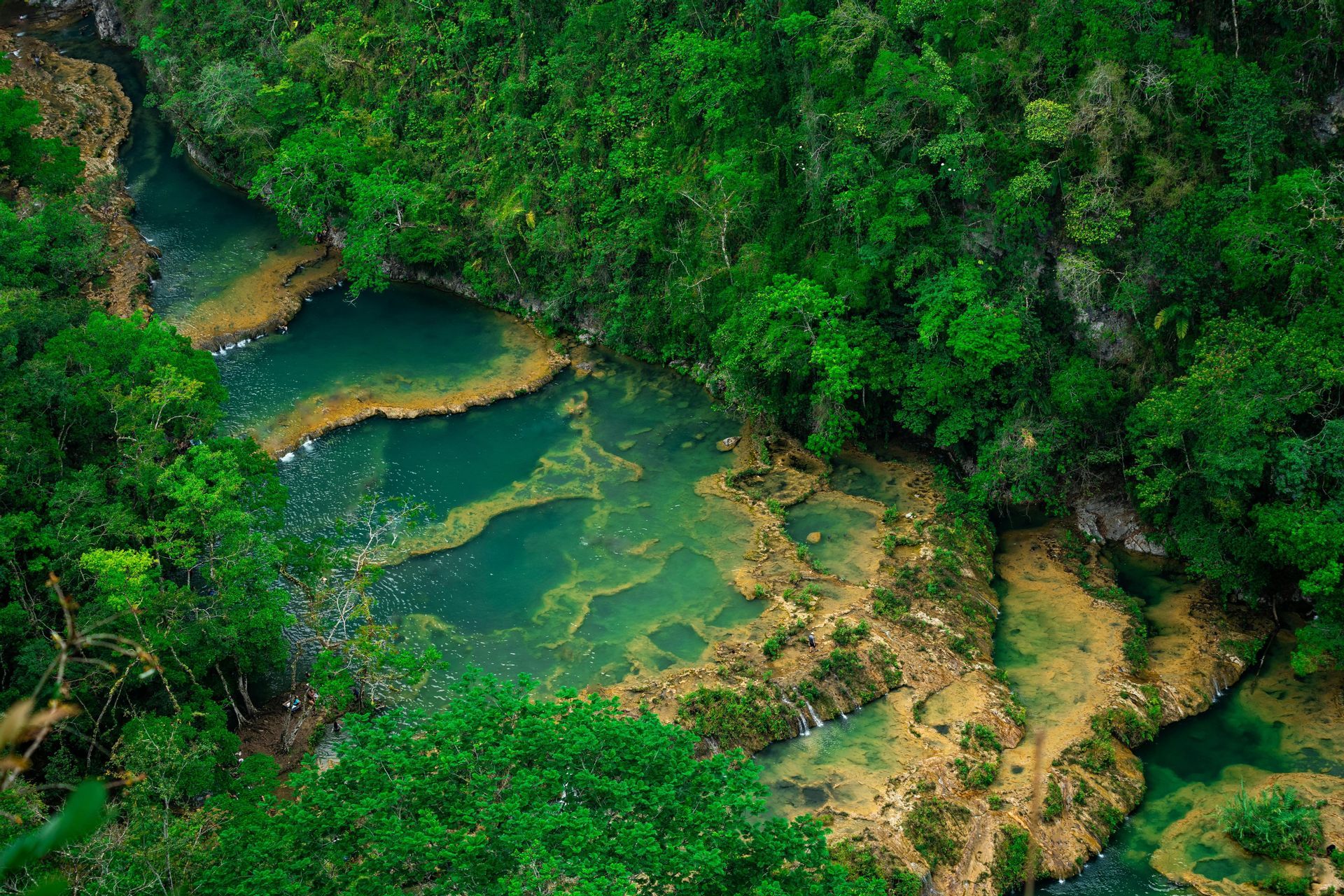 Vue plongeante sur des piscines turquoise en terrasses formant une rivière qui traverse une jungle verte dense.