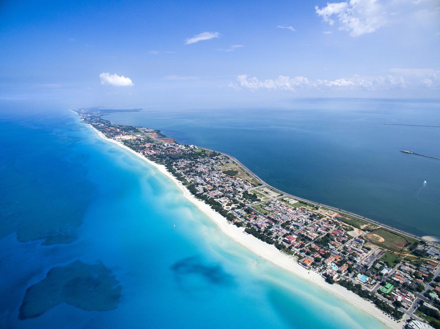 Una vista aérea de una larga península con una playa de arena blanca, un pueblo costero y aguas contrastantes de color turquesa y azul profundo.