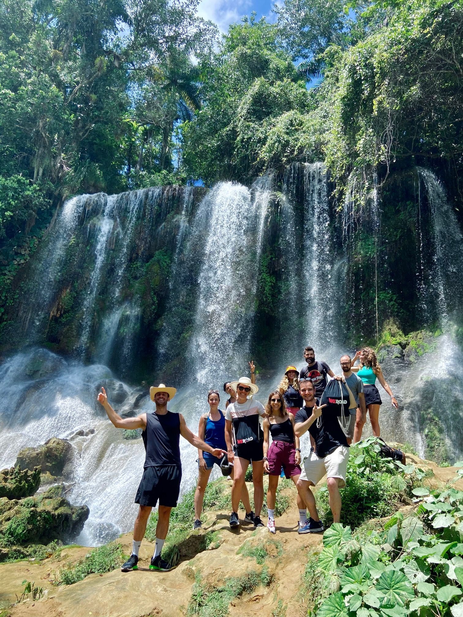 Eine WeRoad-Gruppe posiert für ein Foto auf einem Felsvorsprung vor einem großen, kaskadierenden Wasserfall in einem üppigen Wald.