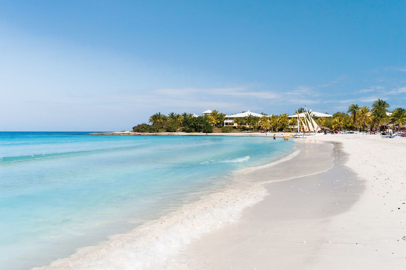 Una spiaggia di sabbia bianca con acque turchesi che lambiscono la riva. Sullo sfondo si ergono palme, barche a vela ed edifici di resort.