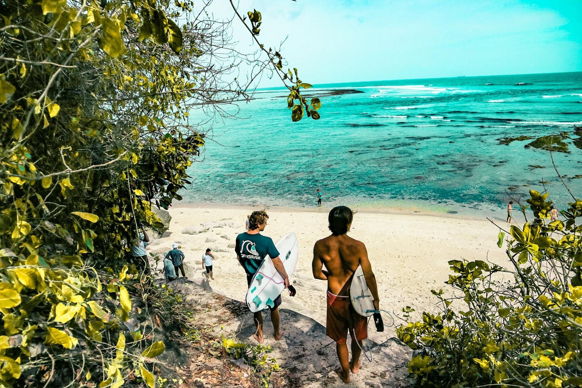 Dos surfistas con sus tablas caminan por un sendero a través de la vegetación hacia una playa de arena y un mar turquesa.