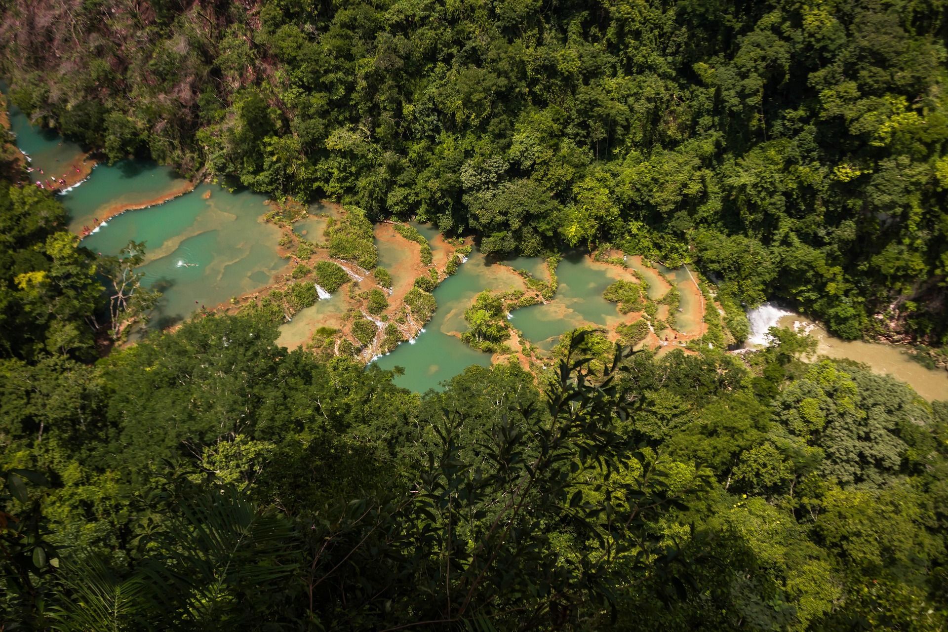 Vista aérea de pozas escalonadas turquesas en un río que serpentea a través de una densa selva verde, con gente visible sobre las rocas.