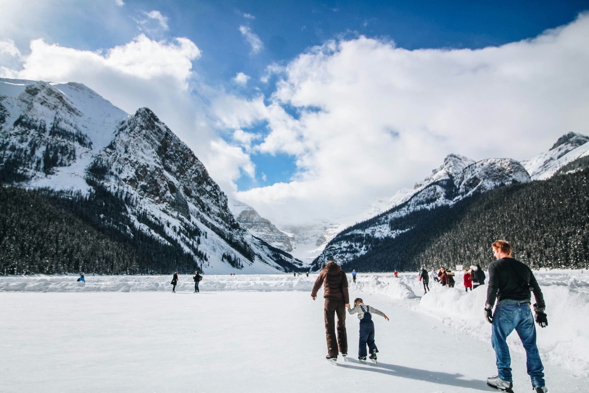 Menschen beim Eislaufen auf einem weiten zugefrorenen See, umgeben von hoch aufragenden schneebedeckten Bergen unter einem teils bewölkten Himmel.