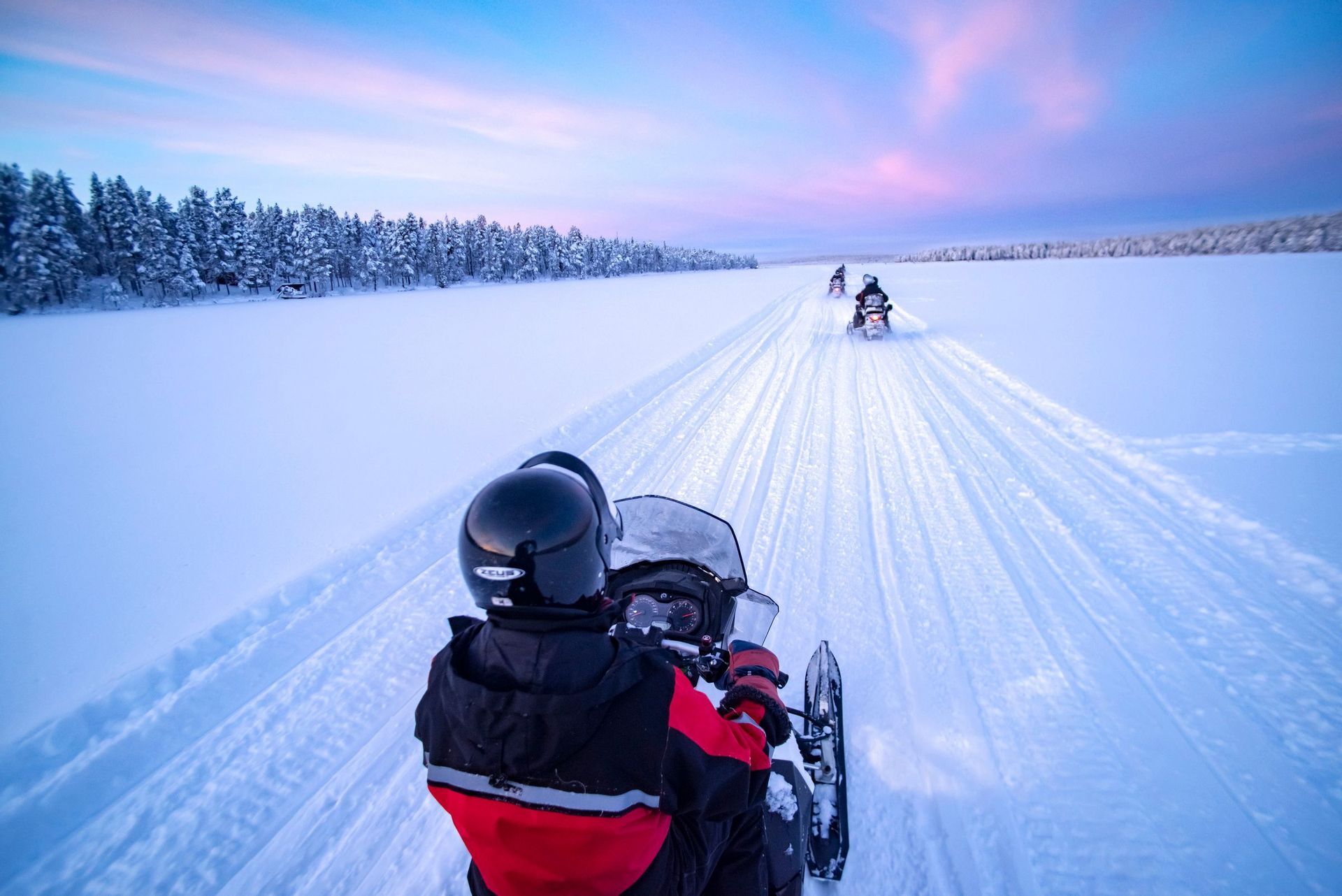 Eine WeRoad Gruppenreise: Schneemobilfahrt in einer Reihe durch eine weite Schneelandschaft bei Sonnenuntergang.