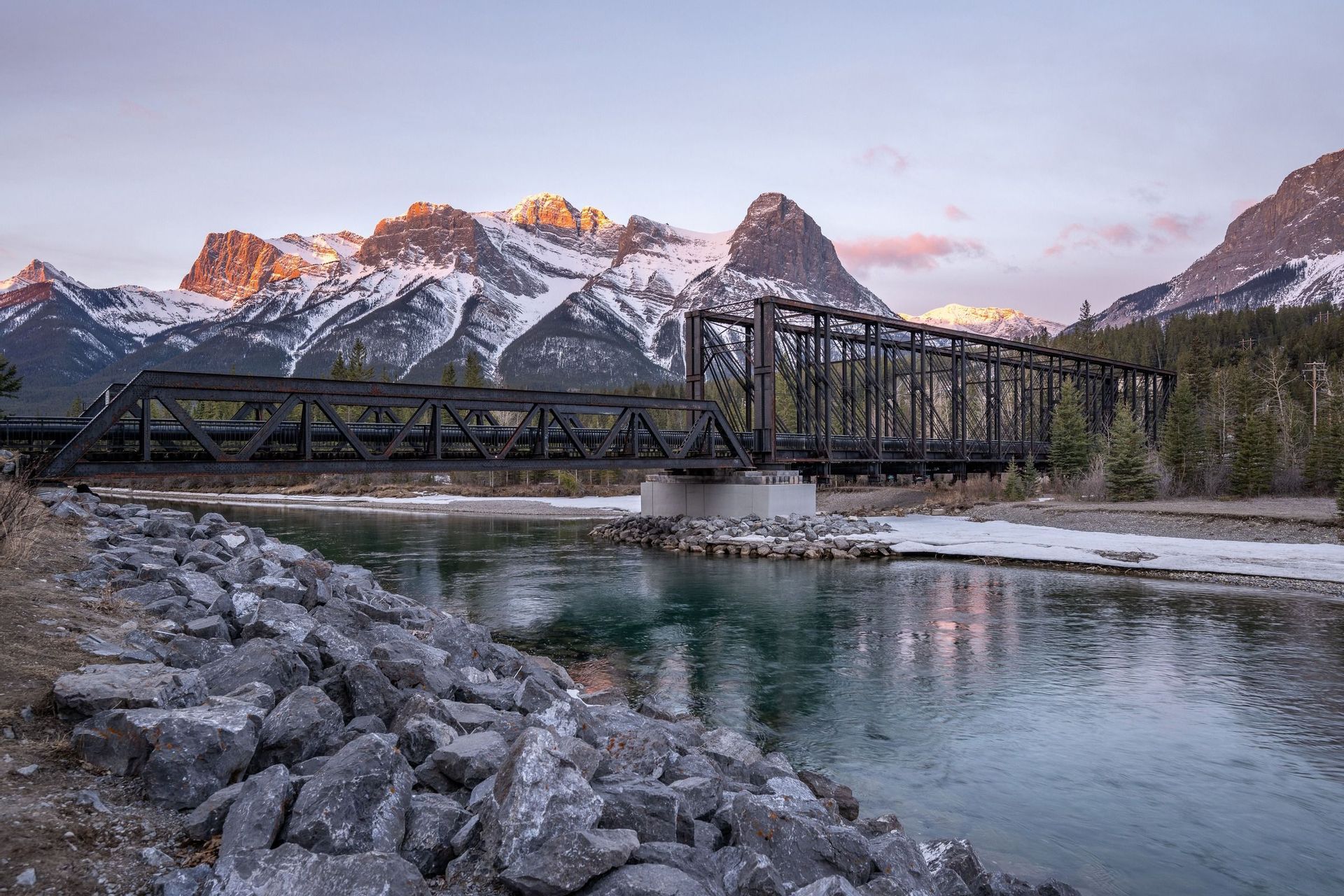 Eine schwarze Eisenbahnbrücke überquert einen Fluss mit felsigem Ufer vor schneebedeckten Bergen mit sonnenbeschienenen Gipfeln in der Dämmerung.