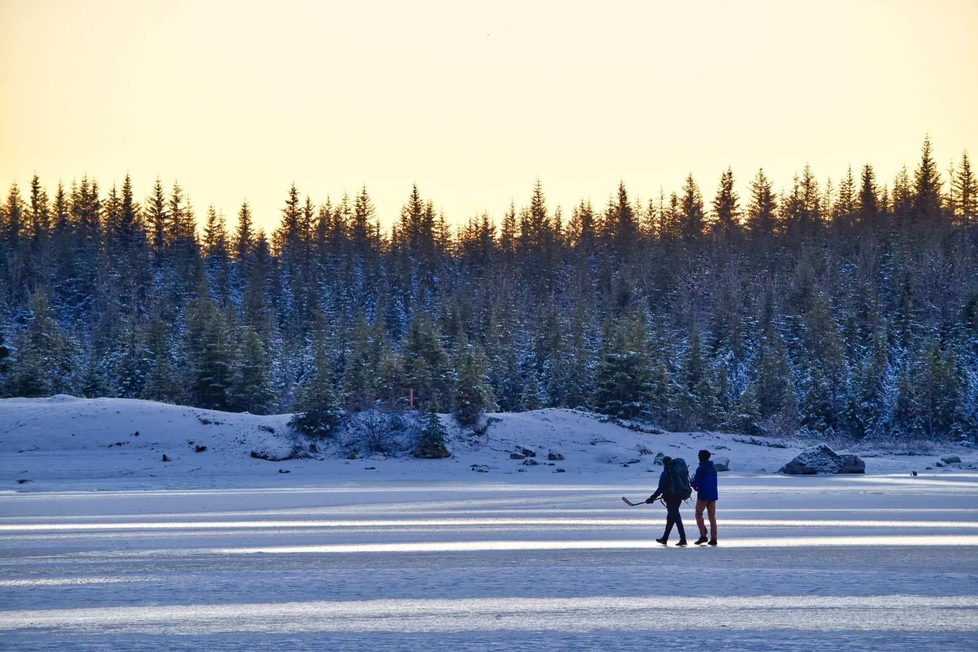 Zwei Personen mit Rucksäcken, eine davon mit einem Hockeyschläger, gehen über einen zugefrorenen See vor einem schneebedeckten Kiefernwald.